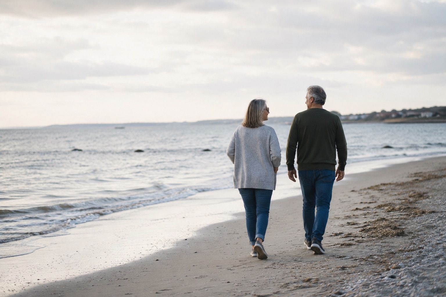 A mature couple walking quietly along the shoreline in soft natural light, reflecting the calm and grounded tone of later-life companionship.
